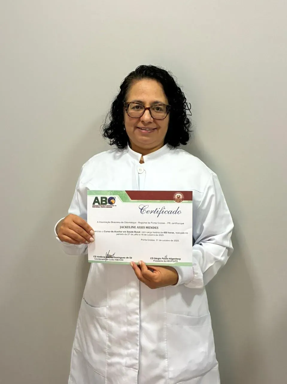 Aluna do Curso ASB de jaleco branco e óculos segurando o certificado com o selo da ABO