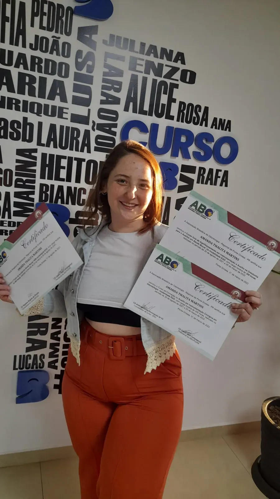 Aluna do Curso ASB sorrindo e segurando dois certificados diante de uma parede com nomes de formandas