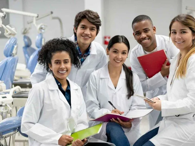 Cinco estudantes de odontologia de jaleco branco, sorrindo, com cadernos nas mãos numa clínica-escola